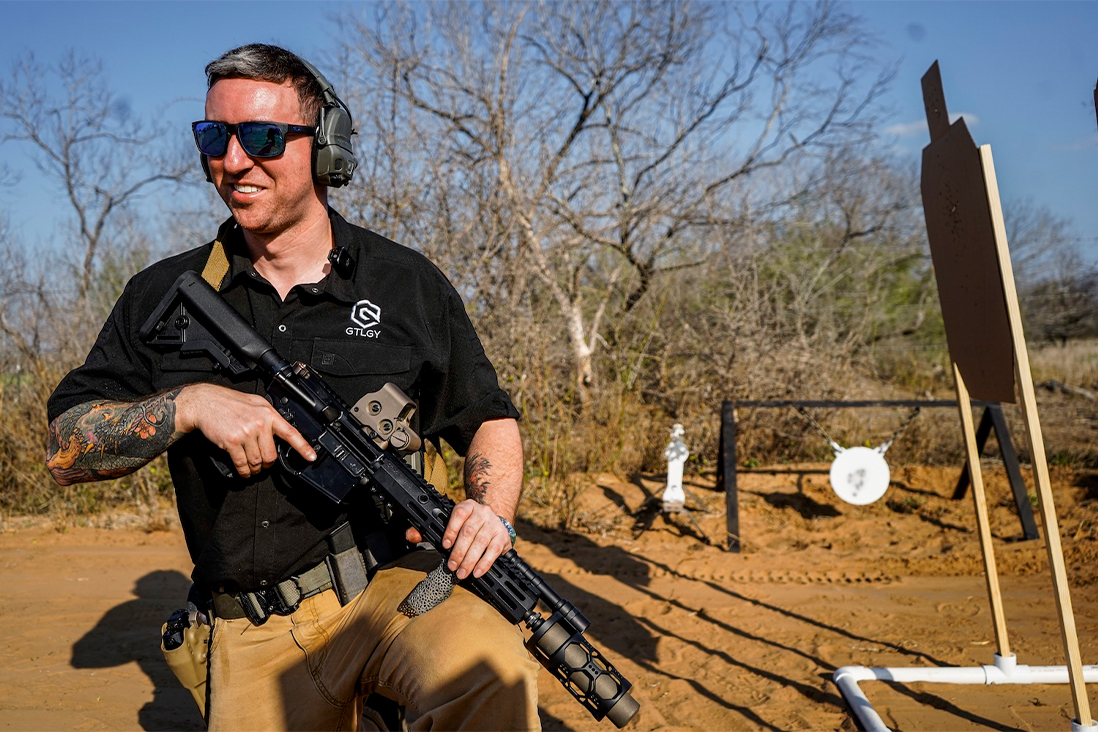 A smiling man wearing sunglasses holds a gun, exuding a confident demeanor.