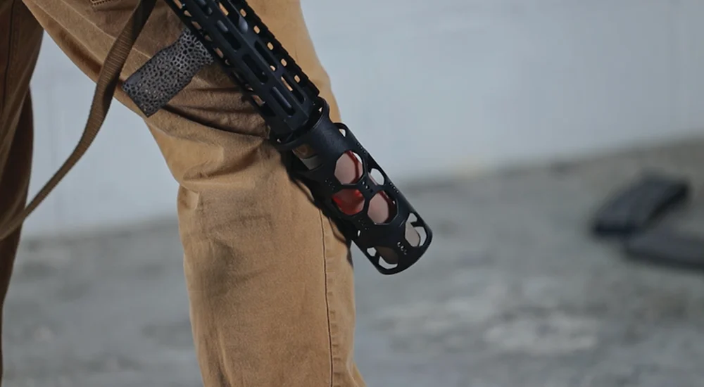 A person stands holding a rifle in their right hand, looking directly at the camera with a serious expression.