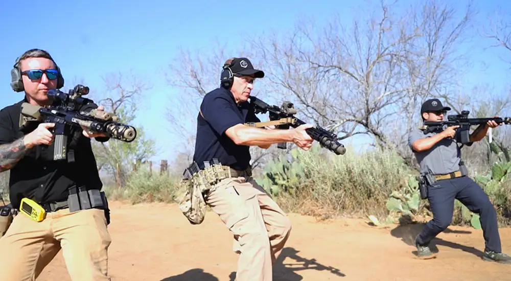 Three men in black shirts and hats are aiming firearms at a target in an outdoor shooting range.