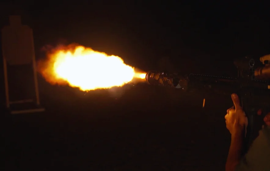 A man aims and fires a gun at night, illuminated by dim light in a dark environment.