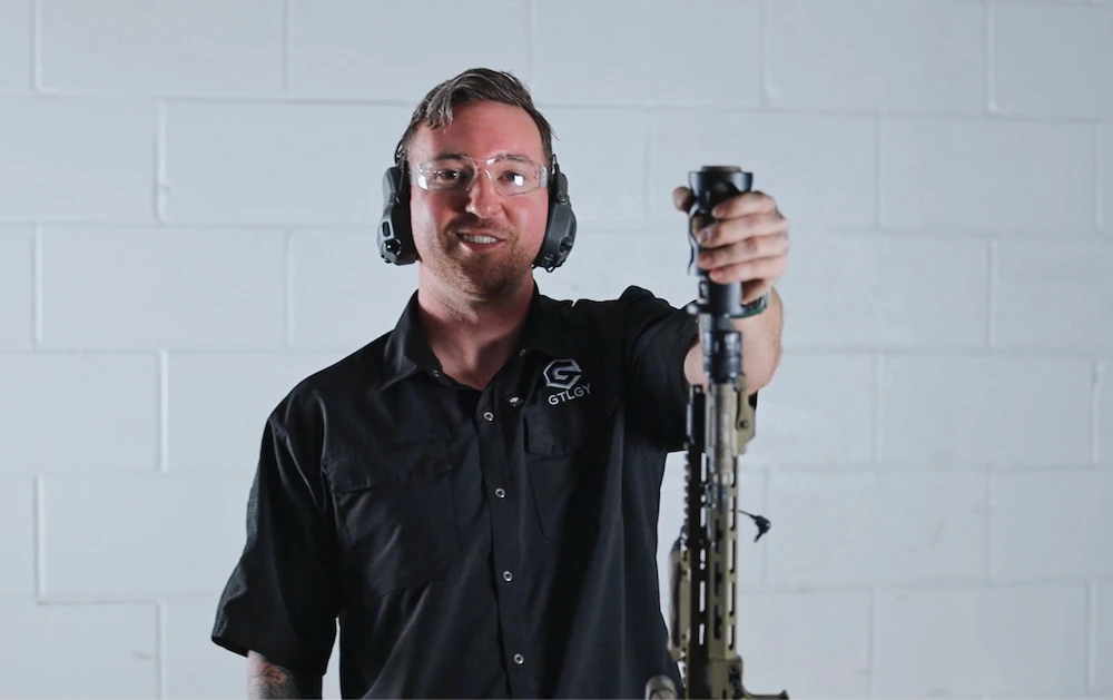 A man stands against a white wall, holding a gun in a ready position.