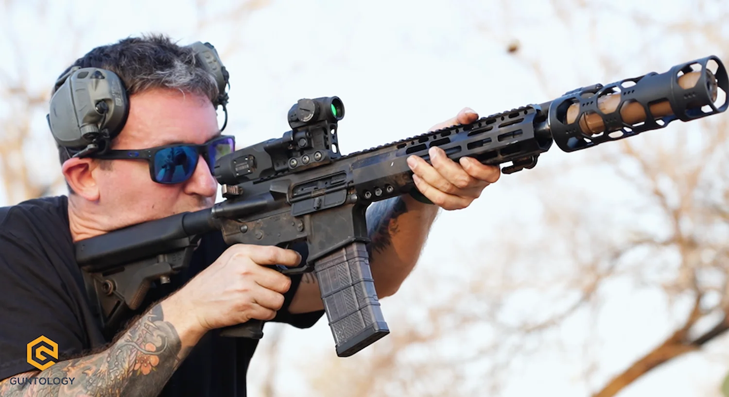 A tattooed man aims an AR-15 rifle, focusing intently on his target.