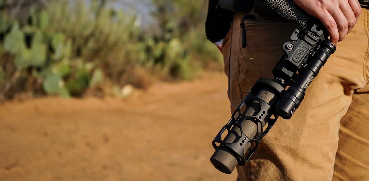 A man grips a rifle while standing on a dirt road, surrounded by a natural landscape.
